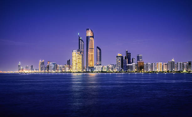 Illuminated nighttime skyline of Abu Dhabi The skyline of Abu Dhabi at sunset, taken from the distance over water. All of the buildings have their lights on, and the large towers in the middle are the Central Market Towers. The sea is very tranquil, and the fading light lingers behind the city. corniche stock pictures, royalty-free photos & images