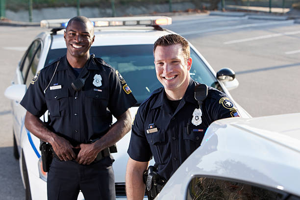 Police officers Multi-ethnic police officers (20s) standing in front of police car. Focus on Caucasian man. police force smiling officer stock pictures, royalty-free photos & images