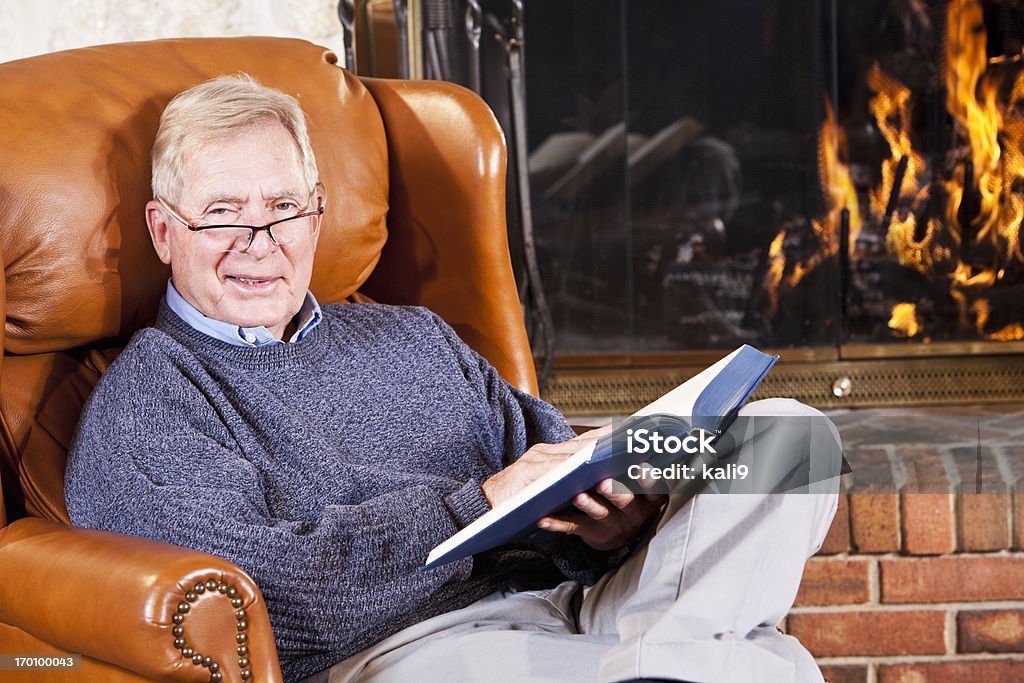 Senior man reading by fireplace Senior man (60s) reading by stone fireplace. Men Stock Photo