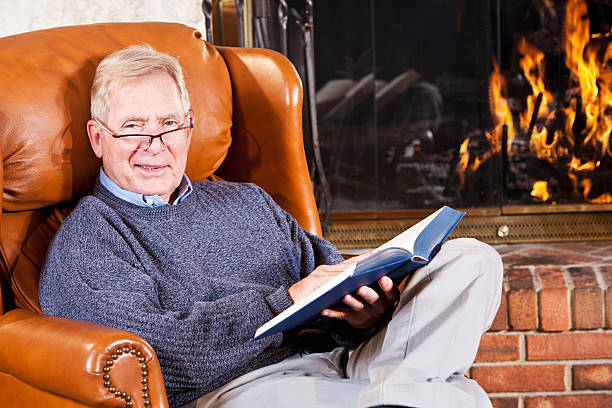 Senior man reading by fireplace Senior man (60s) reading by stone fireplace. fireplace and armchair stock pictures, royalty-free photos & images