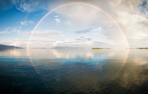 Panorama of a full rainbow reflected in calm water.