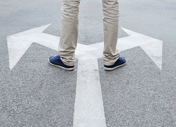 Man standing hesitating to make decision Man standing on arrows painted on asphalt. acute angle photos stock pictures, royalty-free photos & images