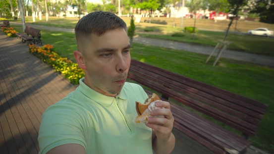 A young man walks through the park and eats a burger. A man films himself while walking along a green alley. 4k