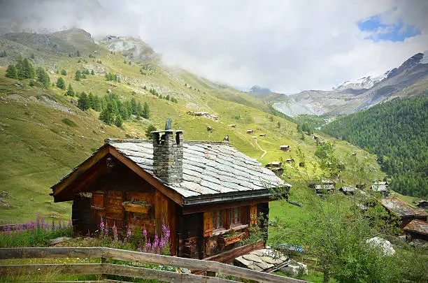 A traditional wooden Swiss chalet with a stone roof sits beside a hiking trail overlooking a small village near Zermatt.