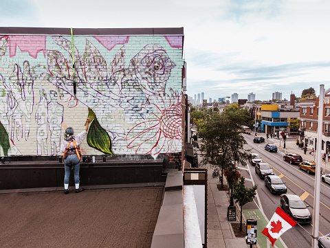 Young Caucasian woman creating outdoor mural on the roof top wall. She is dressed in casual outfit. Exterior of old building in the city of Toronto, Canada..