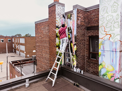 Young Mexican woman creating outdoor mural on the roof top wall. She is dressed in casual outfit. Exterior of old building in the city of Toronto, Canada..