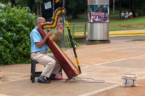 Puerto Iguazu, Argentina - April 3, 2023: Elderly Man sitting and playing Harp Musical Instrument at Hito Tres Fronteras or Triple Frontier, Tri-Border Area between Brazil, Argentina and Paraguay