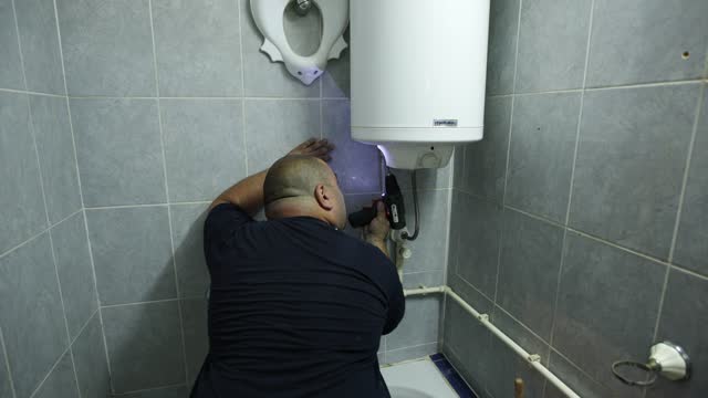 Caucasian male heating engineer using drill machine to repair a boiler at the customer domestic bathroom