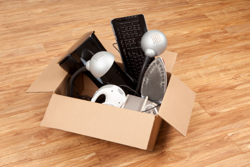 Cardboard box filled with an assortment of household equipment or appliances, ready for moving or a garage sale. Items in the box include a coffee maker,electric mixer, iron, small heater,computer keyboard, and lamps.