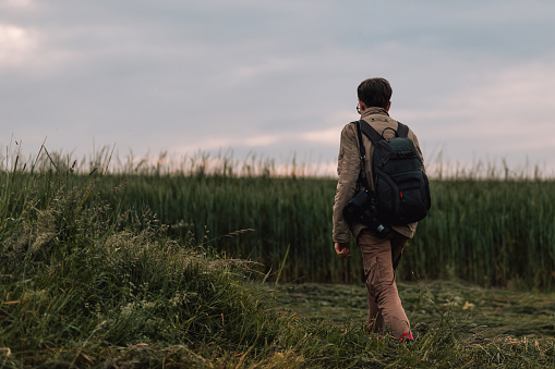 Male portrait, rear view of man with digital camera. A person with a backpack who working photographer walking in the field, traveling concept photography. Lifestyle photo with creative people outdoors. Evening, good weather. Sky, grass, and forest in the background.