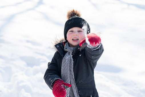 Cute little boy wearing warm winter clothing, a hat, mittens and a winter coat, is laughing as he is throwing a snowball towards the camera. Behind him is a field of cold white snow on a sunny day.