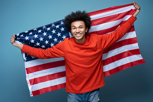 Young happy African American man holding American flag isolated on blue background. Emotional patriot celebration Independence day looking at camera