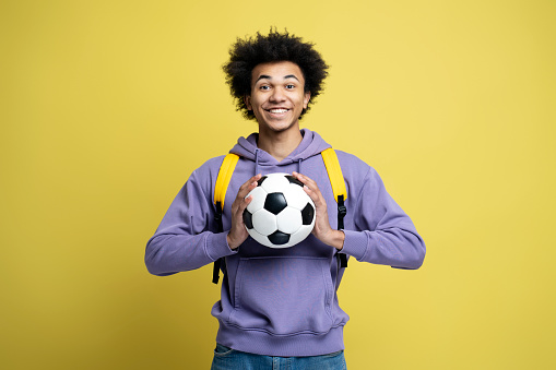 Portrait of young smiling African American man holding ball for soccer game looking at camera isolated on yellow background. Sport, active lifestyle concept