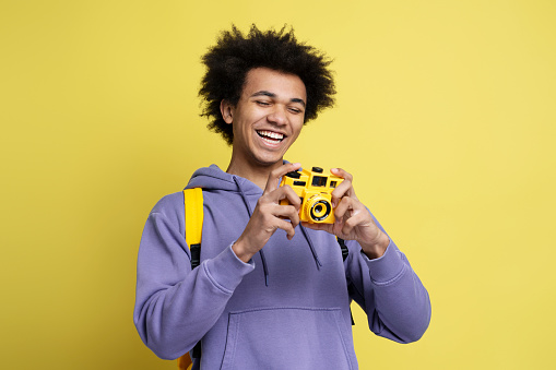 Happy curly haired guy photographer, journalist or traveler with yellow backpack, holding digital camera and smiling looking at camera, before taking photos isolated on yellow background
