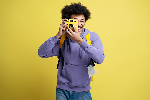 Young smiling African American photographer taking pictures isolated on yellow background. Portrait of happy attractive tourist holding photo camera. Vacation, travel concept