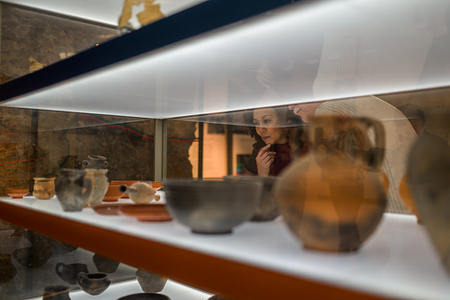 Shot through the glass, close up of a woman's face looking at the exhibited ancient pottery on the shelves, the face of her companion partly visible. Looking away, focus on the earthenware.