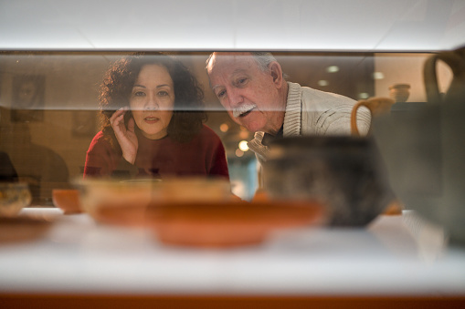 Shot through the glass, close up of the multiracial woman and her Caucasian male friend at the exhibition of ancient earthenware. Both looking away. Blurred pottery in the foreground.