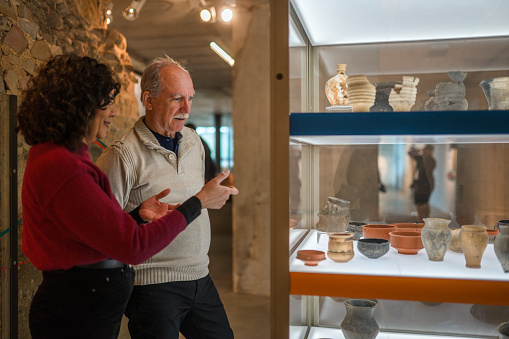 Mature black-haired female curator and her male coworker, both in casual clothes, deciding on the details of an ancient earthenware exhibition. Waist up shot.