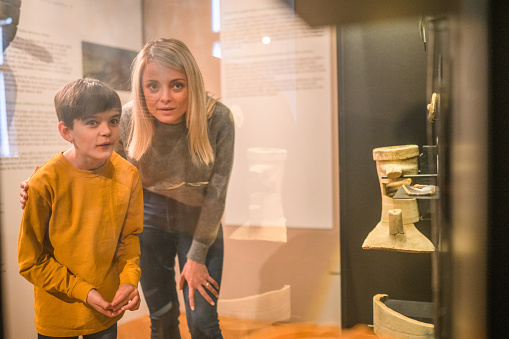 A young Caucasian woman with her  school-aged stepson at a history museum, looking at the objects with admiration. 3/4 length image, shot through the glass, both looking away.