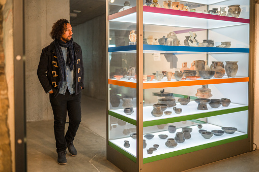Brightly lit glass display shelves and a handsome man next to them, looking at the variety of objects displayed as he walks past it. Full length image.