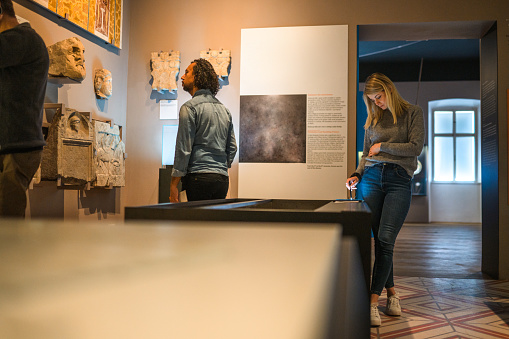 Young Caucasian woman reading through a digital tablet with data of the displayed objects while her biracial husband is checking out the stone remains of an Ancient Roman town. Candid shot over the display cases in the middle of museum gallery.