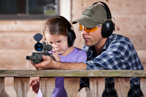 Father teaching daughter how to shoot  rifle.  Main focus on child.