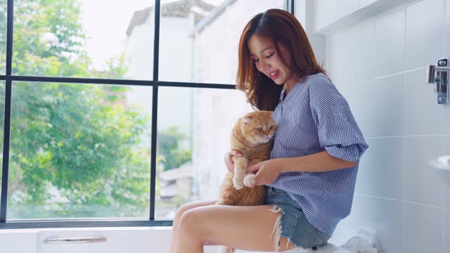 Asian attractive woman playing with domestic cat with happiness at home. Beautiful young female owner sitting on bathtub, feel happy and relax while spend leisure time with her pet animal in bedroom.