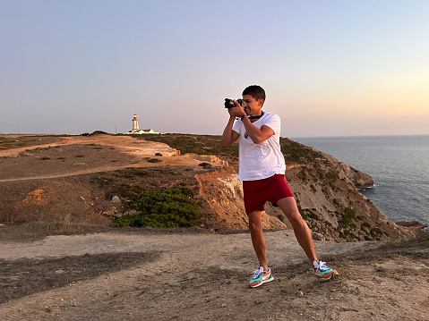 Man takes photos on top of a hill and in the background there is a lighthouse
