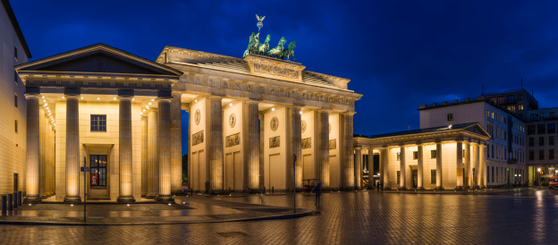 Warm spotlights illuminating the golden stonework of the Brandenburg Gate, iconic symbol of Berlin and Germany reflecting in the cobbles of Pariser Platz under blue dusk skies. ProPhoto RGB profile for maximum color fidelity and gamut.