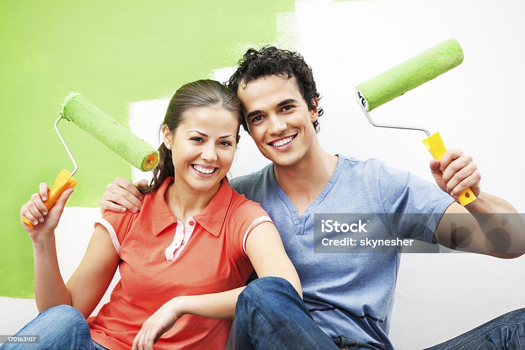 Cheerful couple painting the wall Young couple improving their home. They are embraced holding their paint rollers up and looking at camera.    Adult Stock Photo