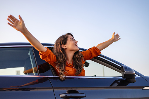Young beautiful women getting new car. she very happy and excited. Smiling female driving vehicle on the road on a bright day. Sticking her head out of the windshield