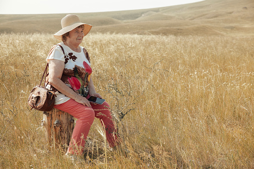 A senior citizen woman in a hat sits with her phone resting in the middle of a wheat field