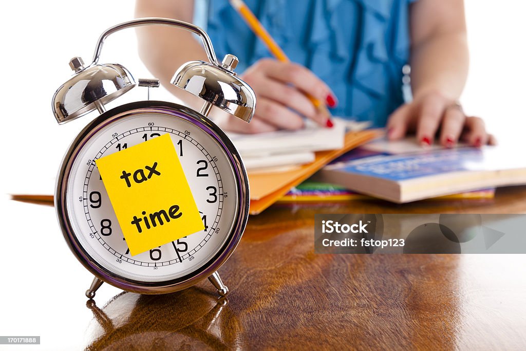 Time:  Alarm clock, income tax time note.  Woman background. Reminder. Time:  Alarm clock with tax time note on front.  Woman in background with paperwork.   Tax Form Stock Photo