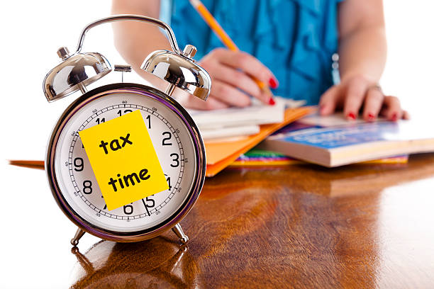 Time:  Alarm clock, income tax time note.  Woman background. Reminder. Time:  Alarm clock with tax time note on front.  Woman in background with paperwork.   tax season photos stock pictures, royalty-free photos & images