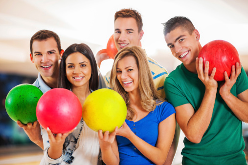 Five friends at the bowling alley are looking at camera and holding the bowling balls.   
