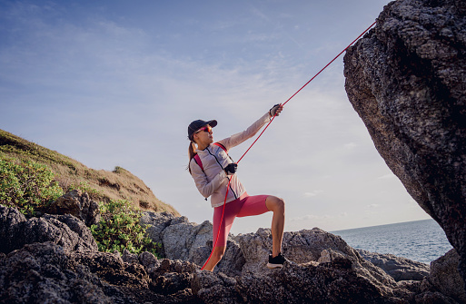 Young woman with a rope climbs to the top in the mountains near the ocean.