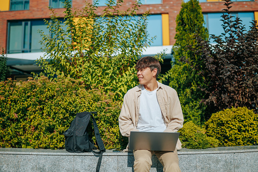 An aspiring student, a young man of Asian Korean descent, engaged in scholarly pursuits, seated on a park bench, intently studying on his laptop in the midst of nature's tranquility.