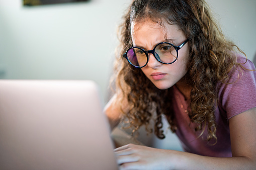 Close-up of serious teenage girl whit eyeglasses looking at laptop while sitting at home