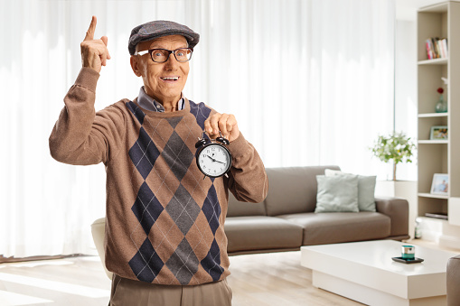 Elderly man holding an alarm clock and pointing up at home in a living room
