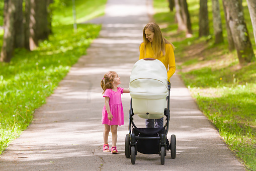 Smiling young adult mother and happy little daughter pushing white baby stroller and walking at town park in warm sunny summer day. Spending time together. Enjoying stroll. Two child mom. Front view.