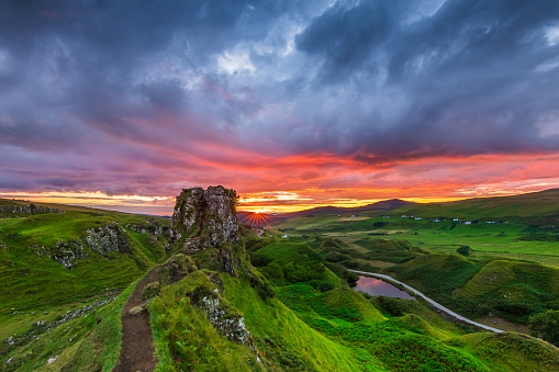 Landscape of an evening mood with sunset in Scotland. Isle of Skye in Scotland in summer. Sun star on the horizon next to Castle Ewen rock. Colorful clouds in the sky. Green meadows and hills