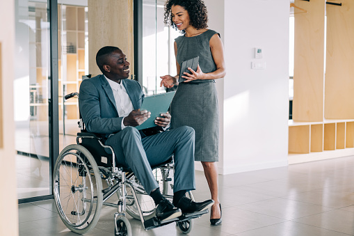 Shot of an african-american businessman in wheelchair having cheerful conversation with his female colleague in the office. Photo of elegant businesswoman using digital tablet while sharing ideas with her disabled colleague.