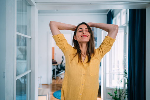 Shot of a beautiful young woman standing with arms behind her head and dreaming with closed eyes at workplace.