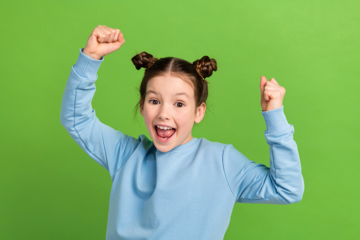 Photo of positive excited schoolgirl with buns hairdo dressed blue pullover raising hands up yell isolated on green color background.