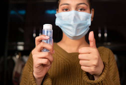 Portrait of young woman wearing surgical protective face mask and holding the hand sanitizer bottle in hand and showing thumbs up to the camera.