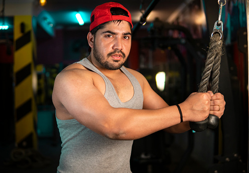Serious sporty muscular young Indian man training arms triceps on the cable machine in the gym. He is looking at the camera with a blank expression. Health and fitness gym concept