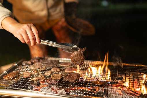 A close up of a Japanese woman grilling meat on a barbecue at camp at night.