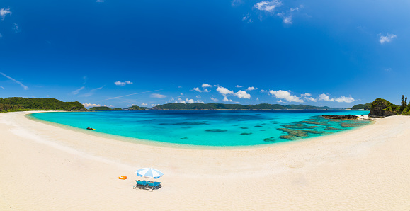 A stunning empty beach on Zamami island in the Kerama island chain of Okinawa with no people. A set of deck chairs and sun umbrella are on the beach. The reef is visible through the clear water.