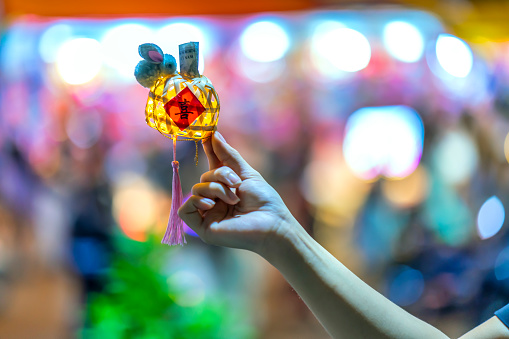 Ho Chi Minh City, Vietnam - September 22nd, 2023: Girl's hand holding a yellow spherical shape lantern for mid-autumn festival decoration on background night lights. The Chinese word means fortune.