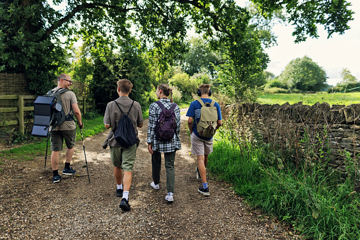 Father and three teenage kids are hiking in the Cotswolds fields in Gloucestershire, United Kingdom. They are walking on a country road. Father is wearing a backpack with a solar panel.\nShot with Canon R5